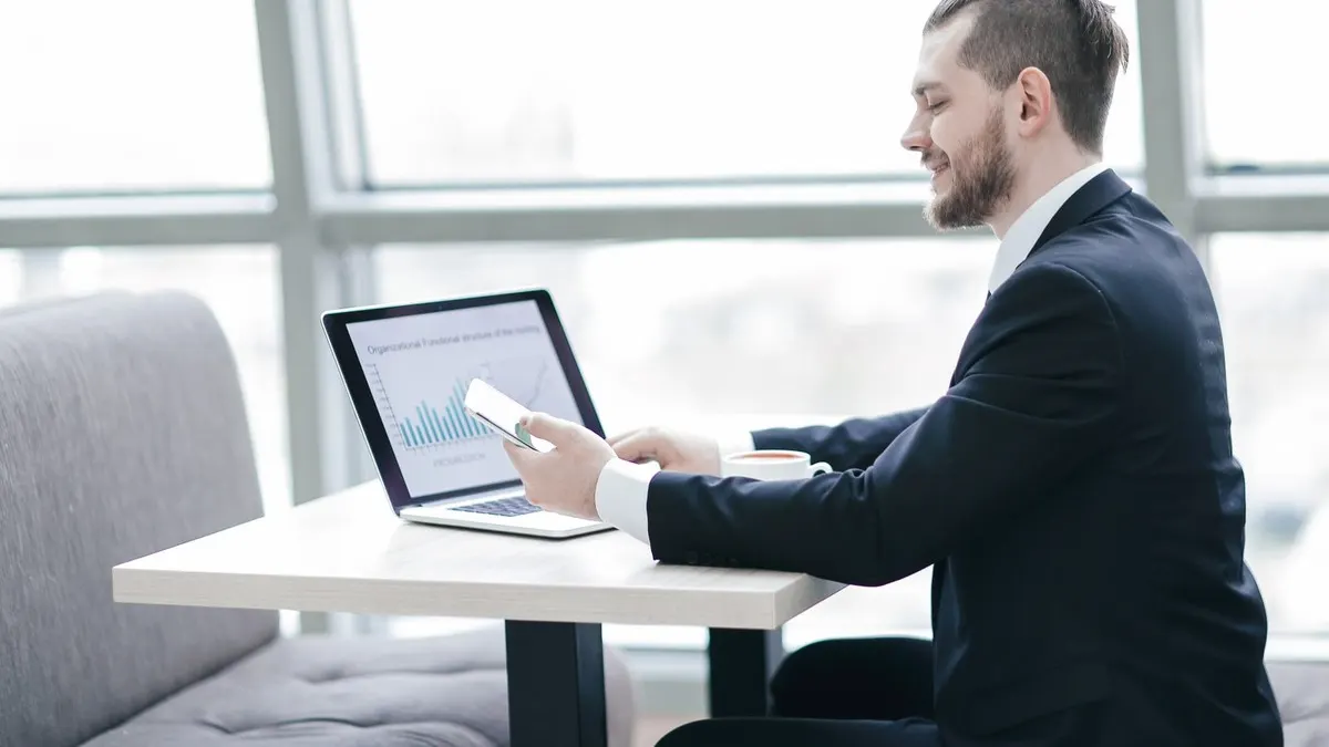 A smiling professional checks his phone while working at a laptop by the tall windows of a bright modern office.