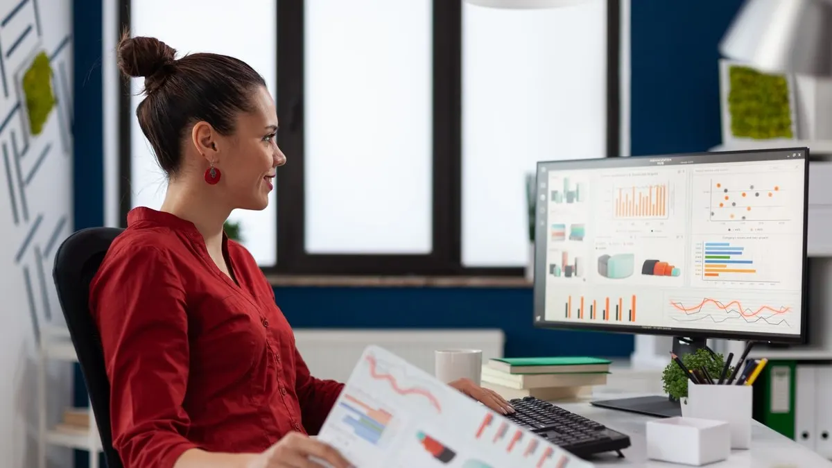 A smiling woman in a red shirt comparing printed charts with the diagrams on her computer screen in a bright office