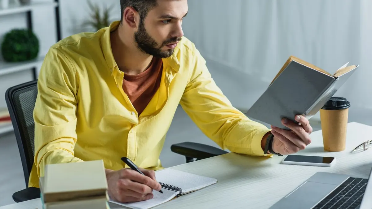 A man in a yellow shirt reads a book and writes notes at a desk beside his laptop in a bright office.