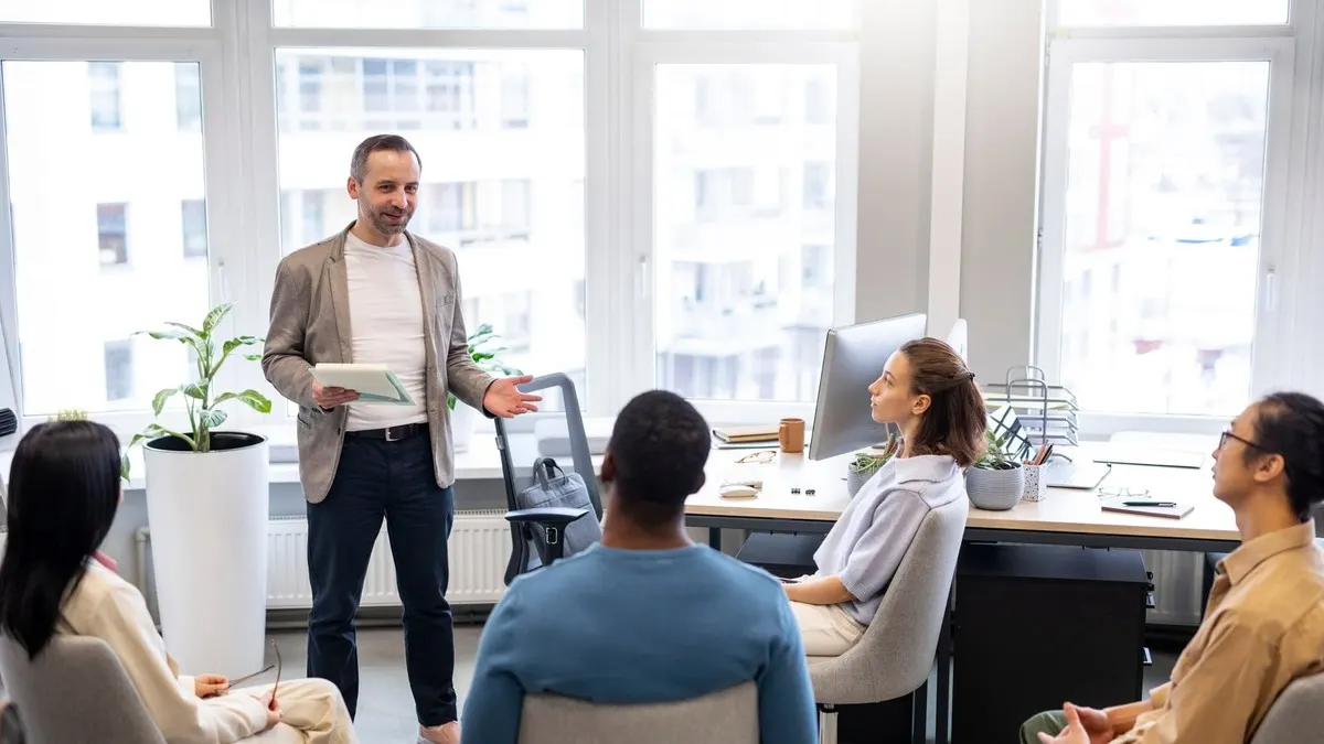 A trainer stands and teaches a small group of seated adult learners in a bright classroom with tall windows.