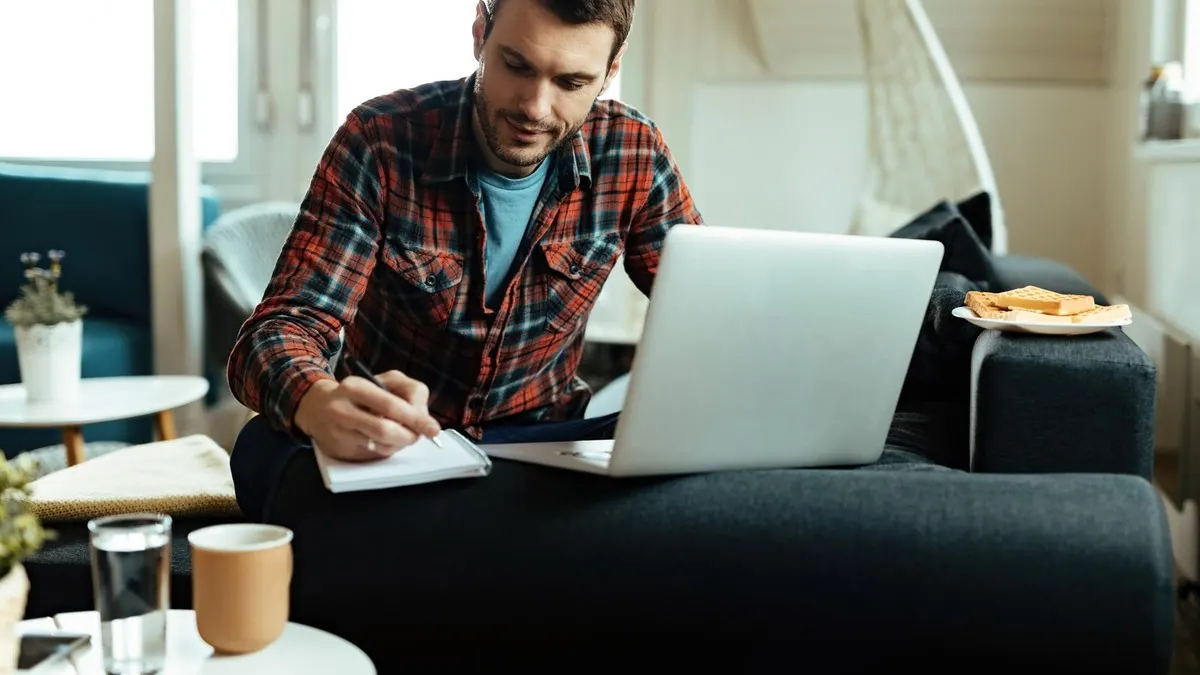 A man in a plaid shirt writes notes beside his laptop while planning at home in a bright living room.