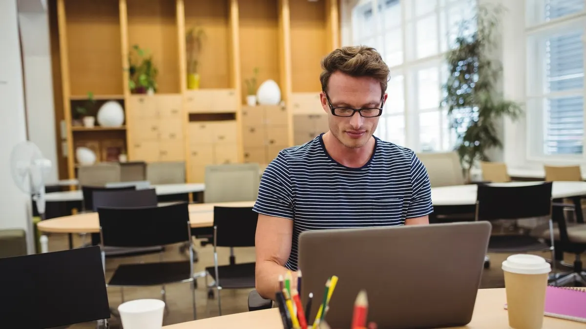 A man in glasses and a striped T-shirt works on a laptop in a bright open office