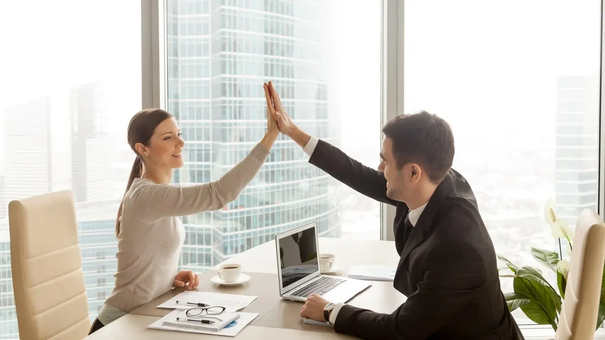 Two colleagues giving each other a high five across their desks in a bright office with city windows