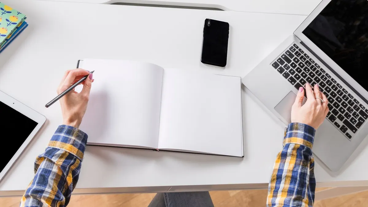 Seen from above, a person writes in an open blank notebook while typing on a laptop at a white desk