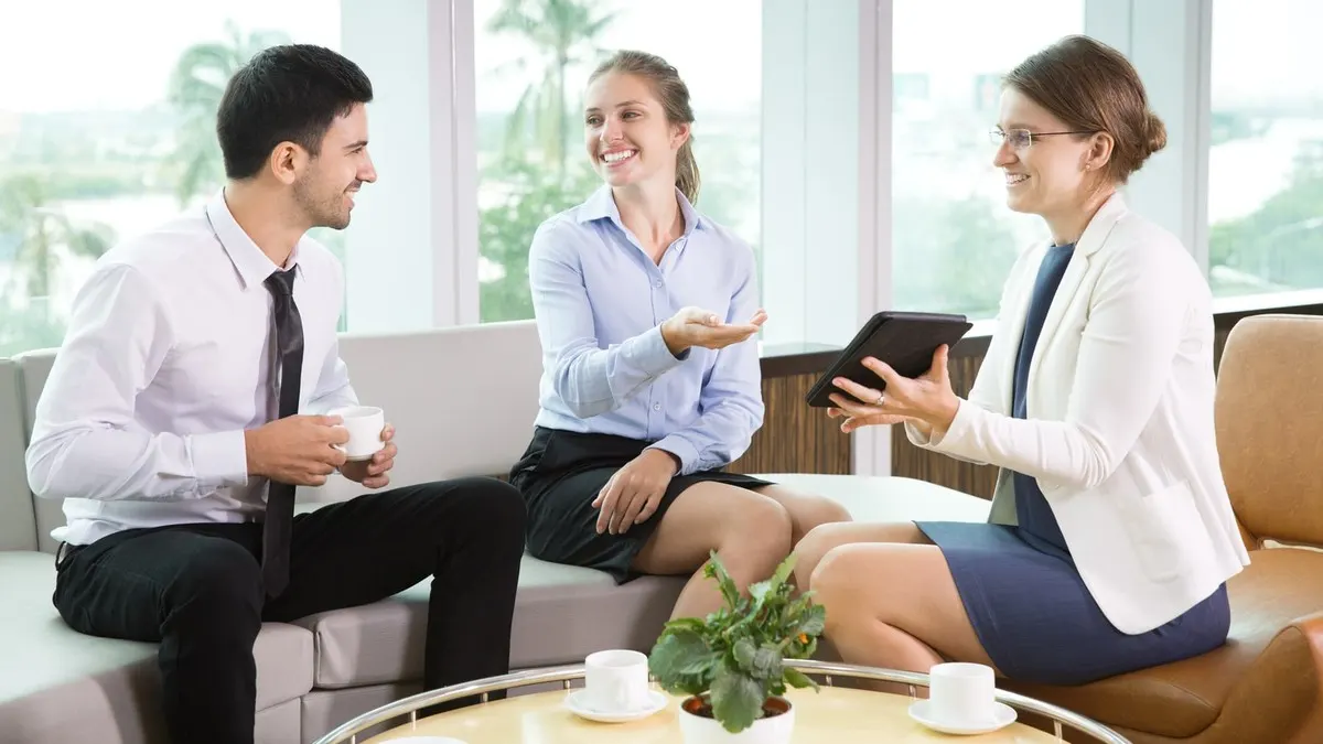 Three colleagues chatting over coffee on sofas in a bright office lounge