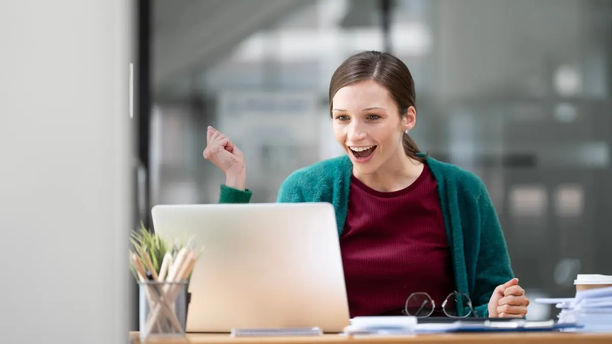 A young woman at her office desk raising a fist in celebration as she reads good news on her laptop