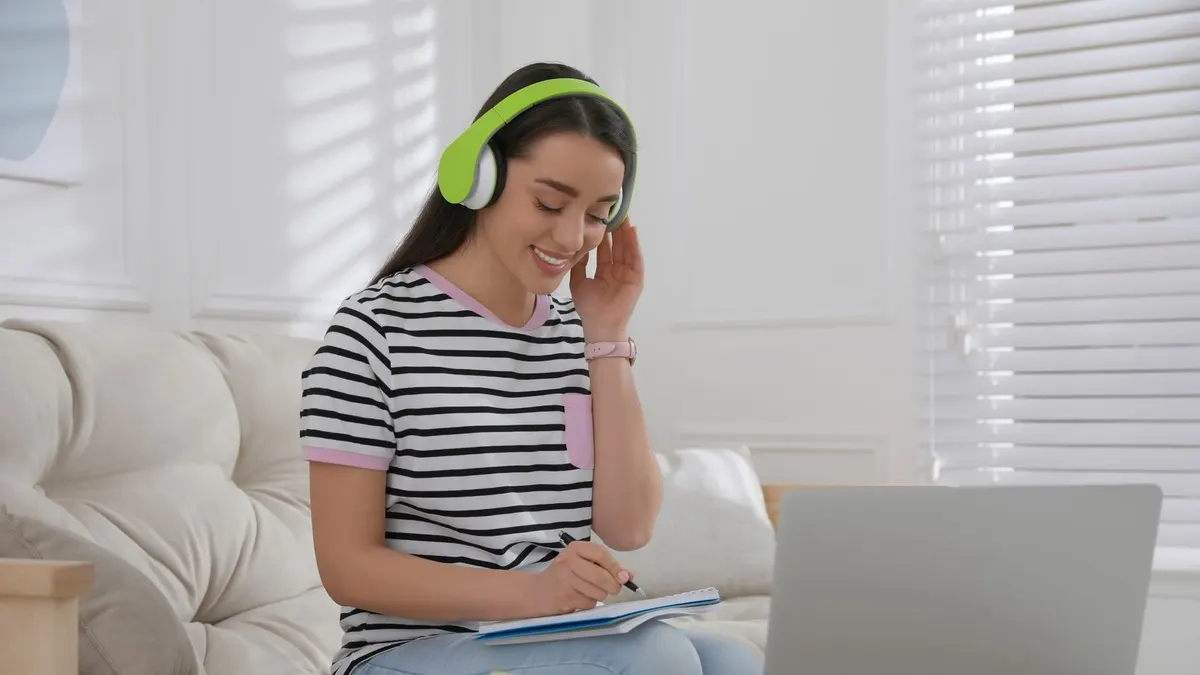 A smiling woman wearing headphones taking notes in front of a laptop in a bright white living room