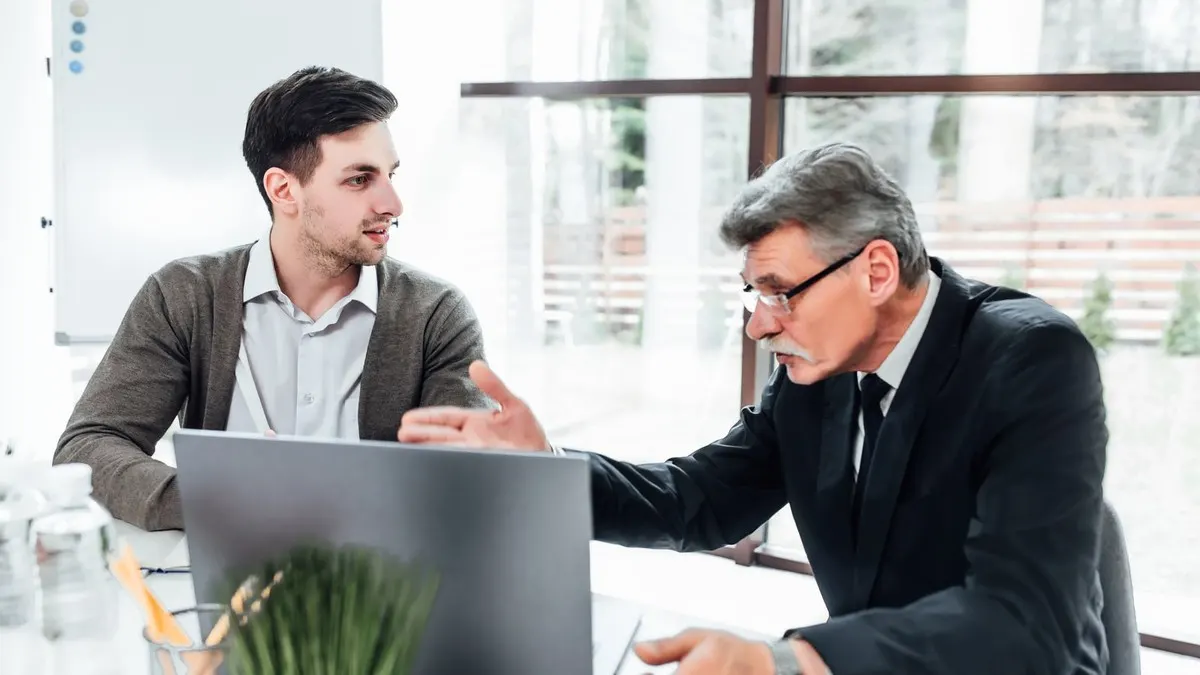 A senior manager in a dark suit explains something at a laptop to a younger colleague beside a bright window.