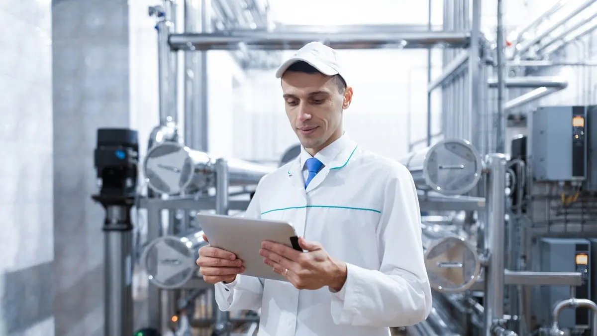 A quality technologist in a white coat and cap checking readings on a tablet beside steel pipes and gauges in a production plant
