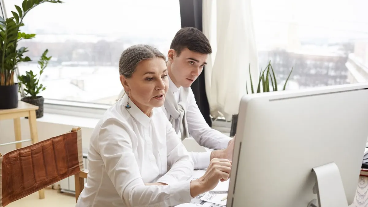 A grey-haired woman points at a desktop monitor while a young colleague leans in to follow, beside a tall bright window.