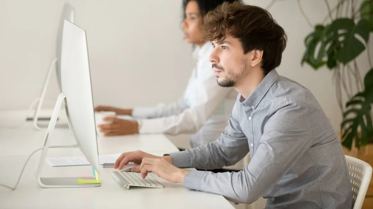 A young man typing at a desktop computer in a bright office with a colleague beside him