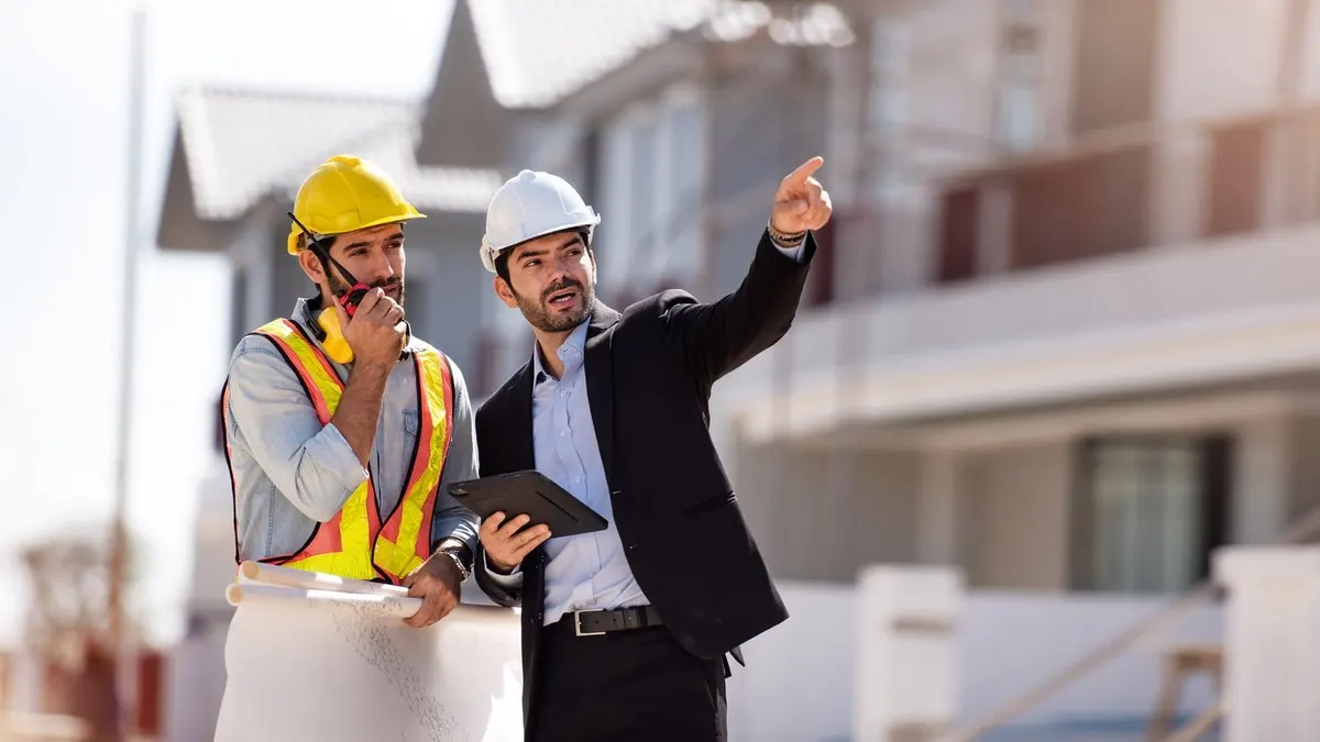 A manager with a tablet points across a housing site while a worker in a hard hat speaks into a radio