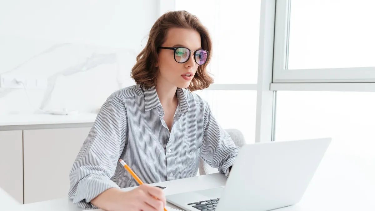 A young woman in glasses takes notes beside her laptop while studying at a table in a bright room.