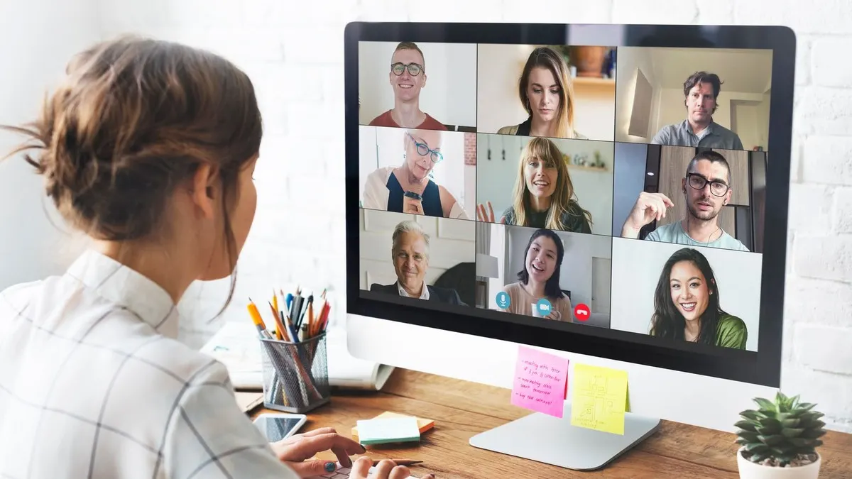 A woman at a wooden desk joins a video call with nine colleagues shown in a grid on her desktop screen.
