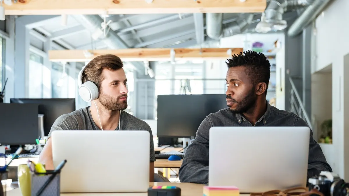 Two developers sitting side by side at their own laptops turning to compare notes in a bright open-plan office