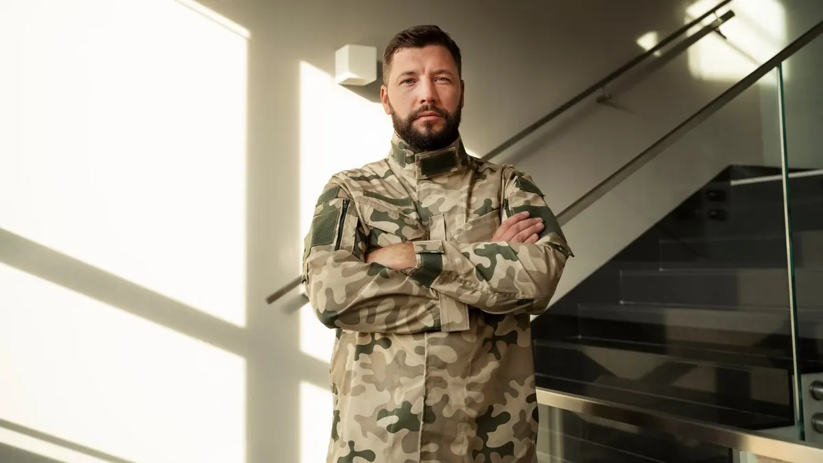 A bearded man in a camouflage uniform standing with arms crossed in a sunlit hallway