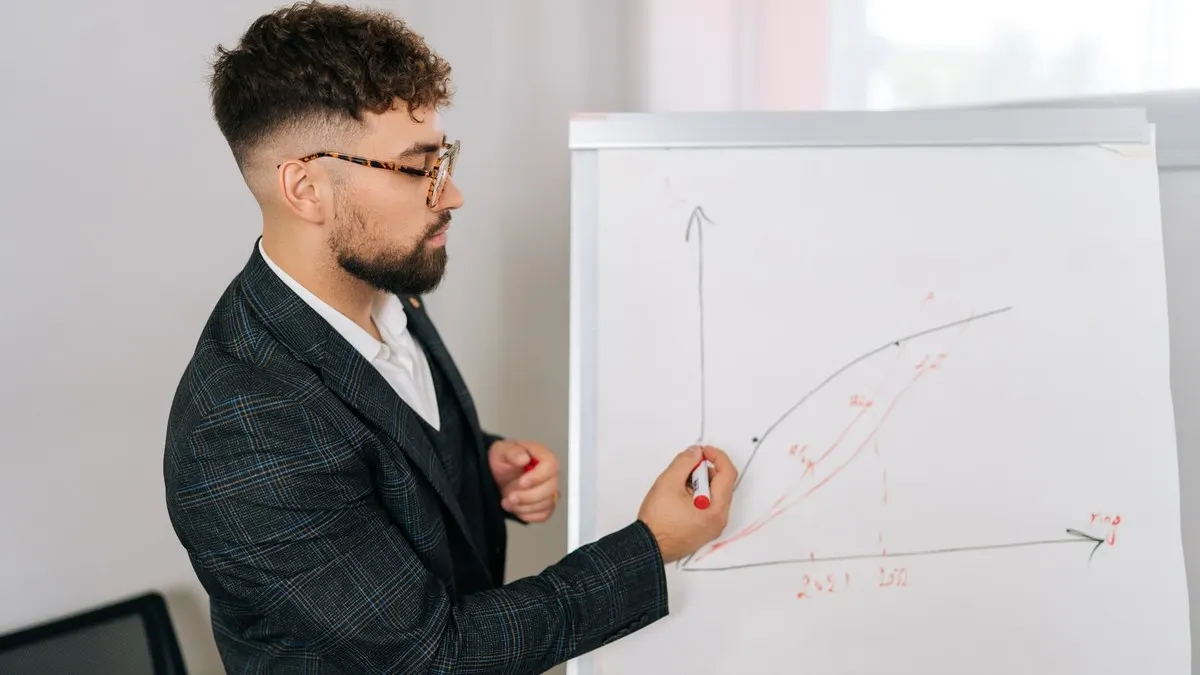 A young businessman in glasses drawing a rising curve on a flipchart in a bright office