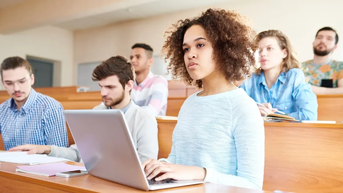 University students listening attentively in a bright lecture hall, one taking notes on a laptop