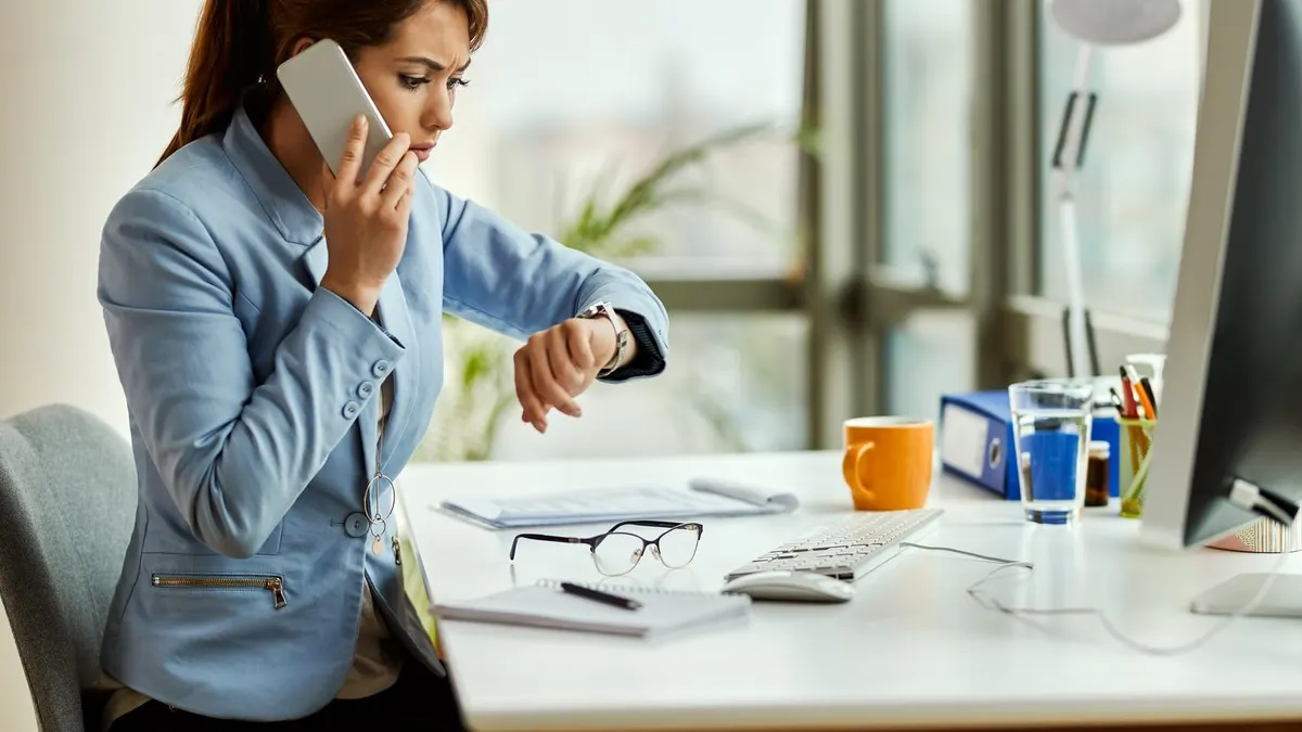 A worried woman in a light blue blazer talks on her phone while checking her wristwatch at a bright office desk.