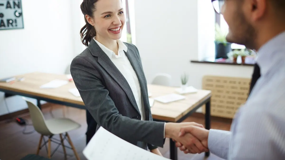 A smiling woman in a grey blazer shakes hands with a man holding a document in a bright office