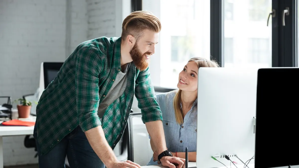 A bearded developer leaning over a desk to talk through code with a colleague at her desktop computer in a bright office