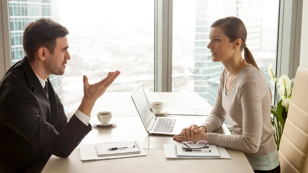 A man gesturing as he argues a point with a woman across a desk by a bright window