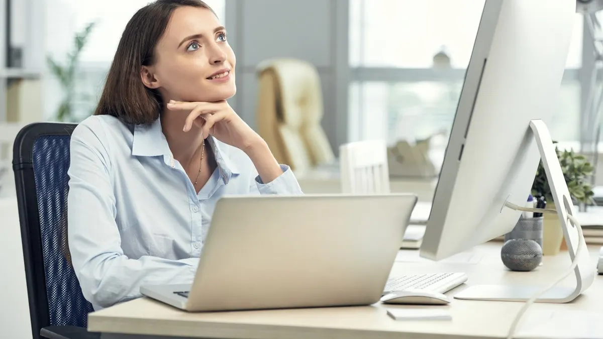 A woman resting her chin on her hand as she thinks through a problem at her laptop and desktop screen in a bright white office