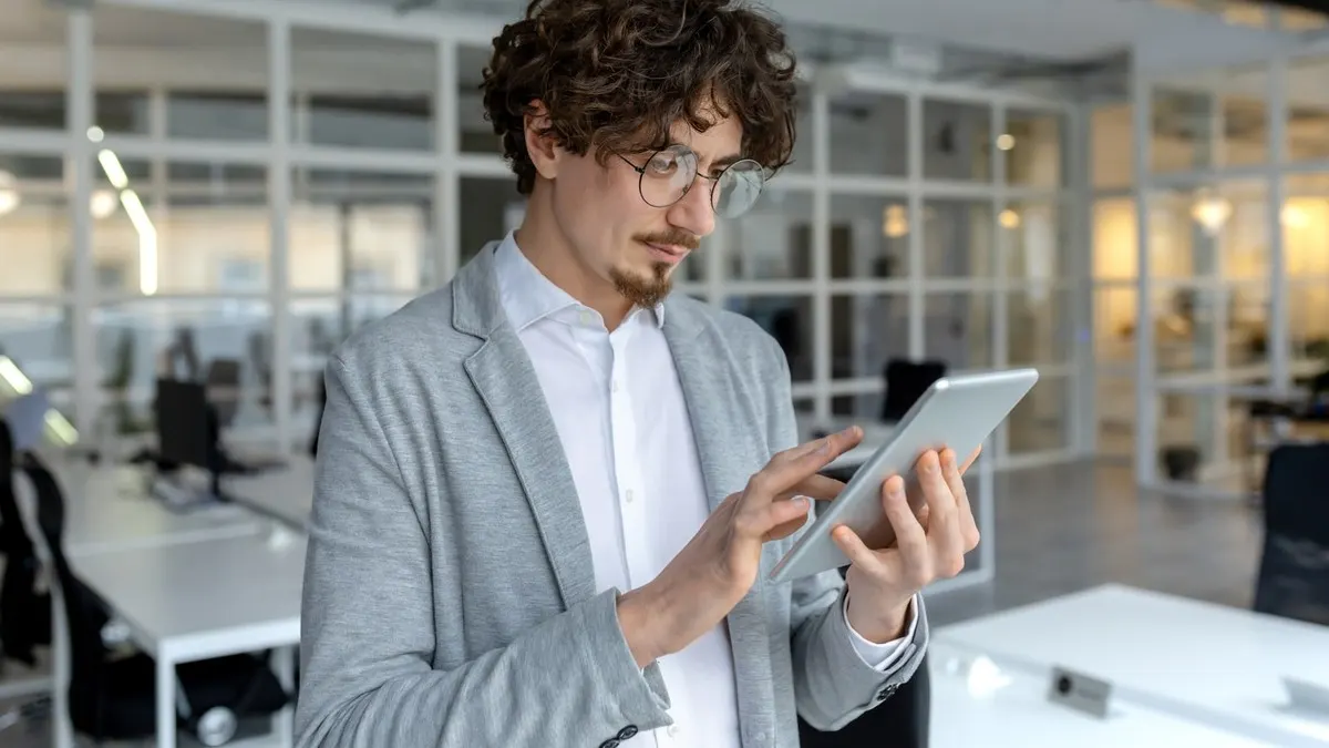 A curly-haired man in glasses and a grey blazer taps on a tablet while standing in a bright open-plan office.