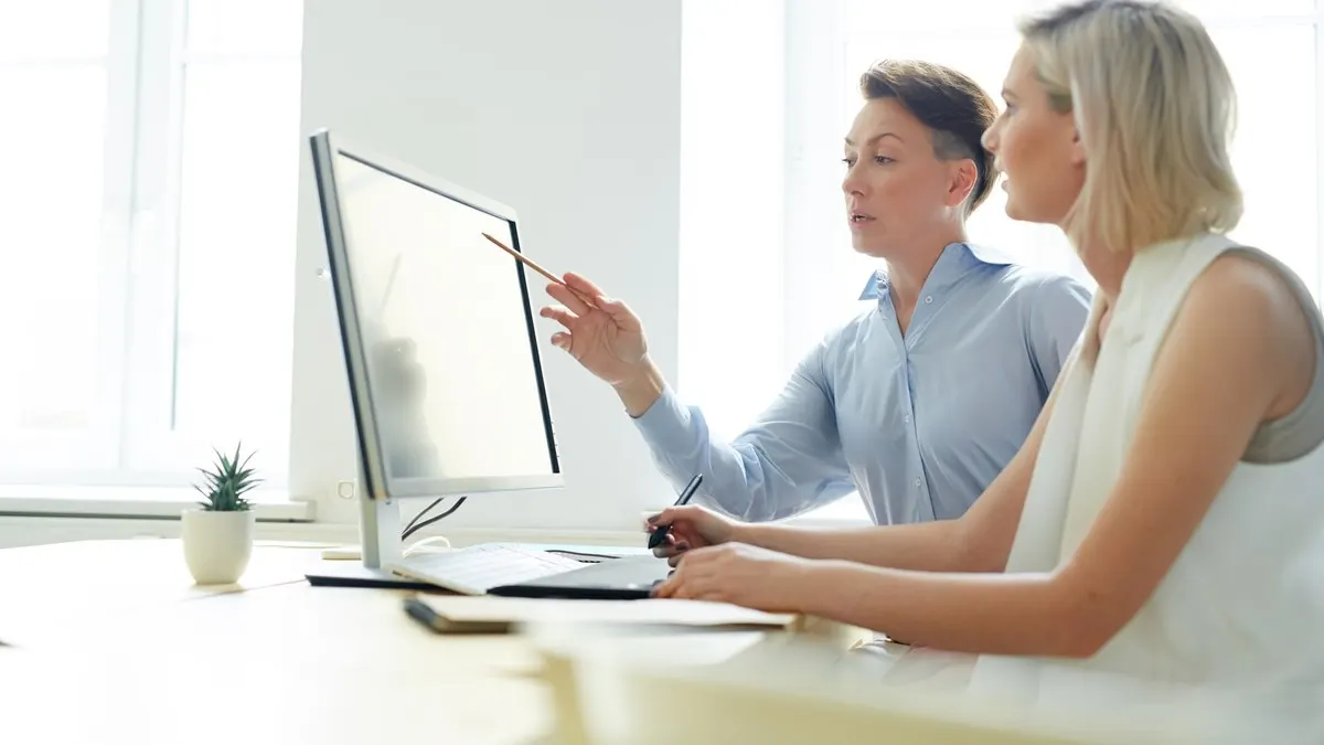 Two women reviewing work on a computer monitor at a desk in a bright white office