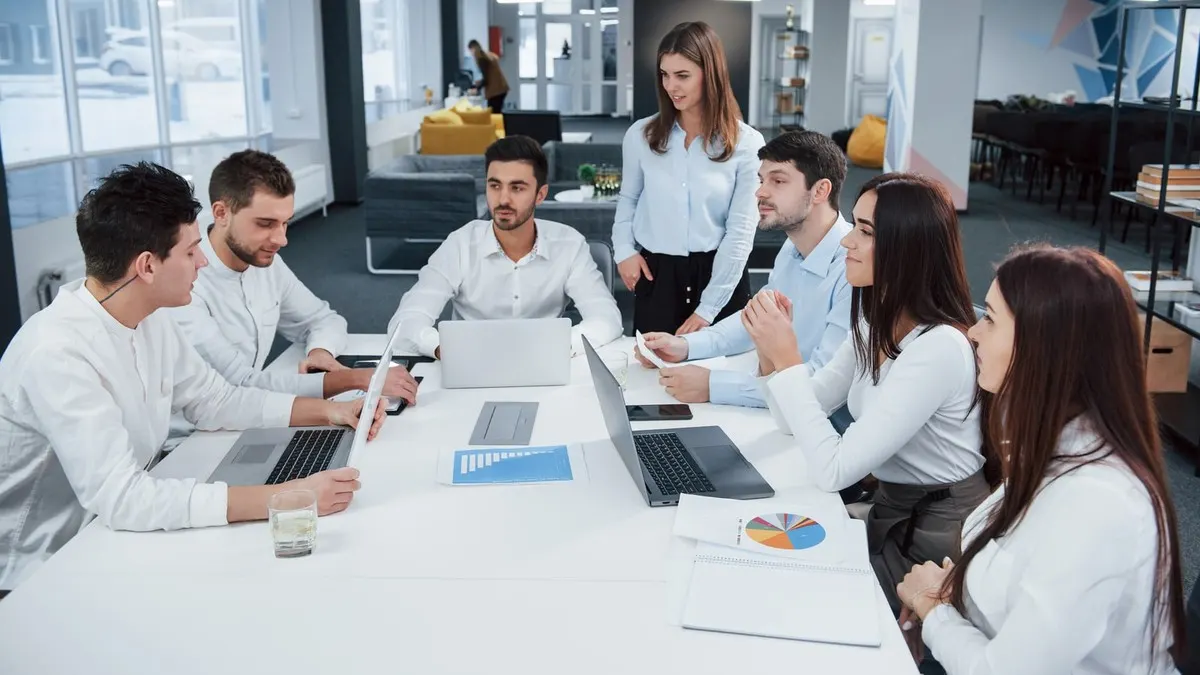 A team of seven colleagues talking around a long white table with laptops in a bright open office