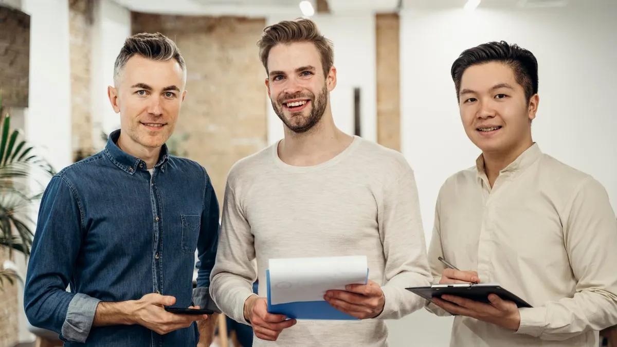 Three smiling young engineers standing side by side with tablets and a clipboard in a bright open office