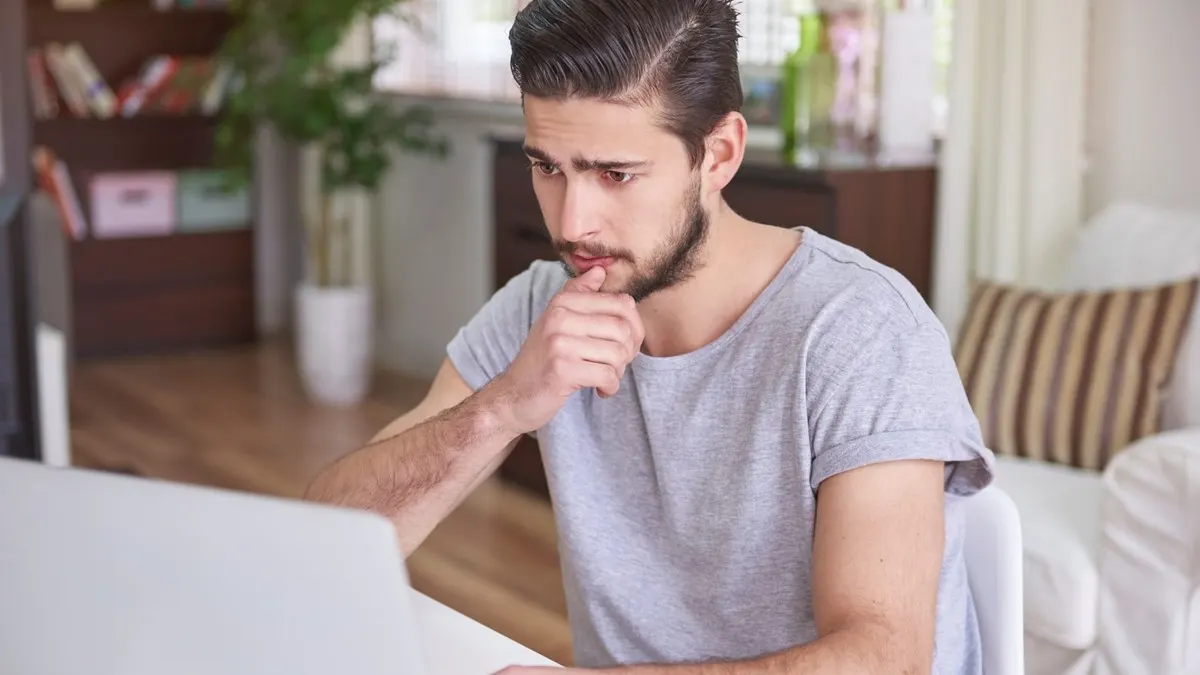 A young man in a grey t-shirt frowns at his laptop with his hand at his chin, unsure of an answer.