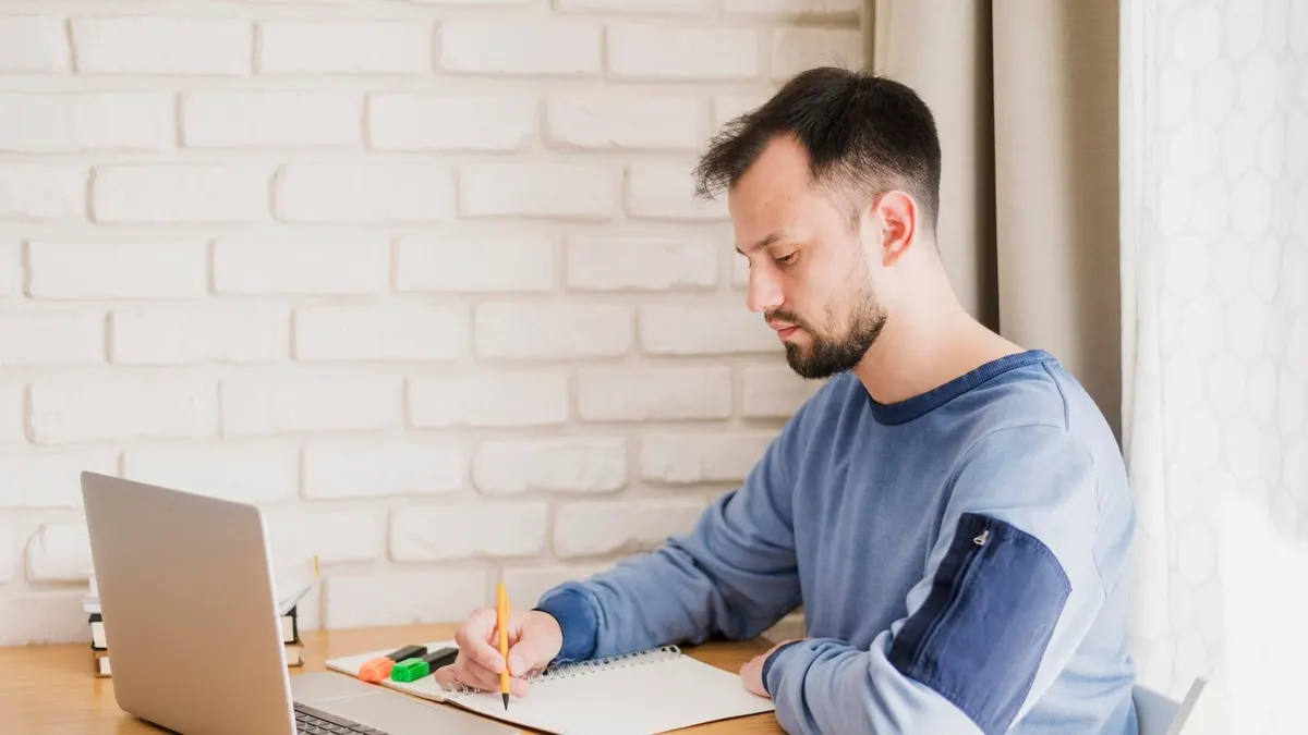 A young man writes notes in a spiral notebook beside his laptop against a white brick wall