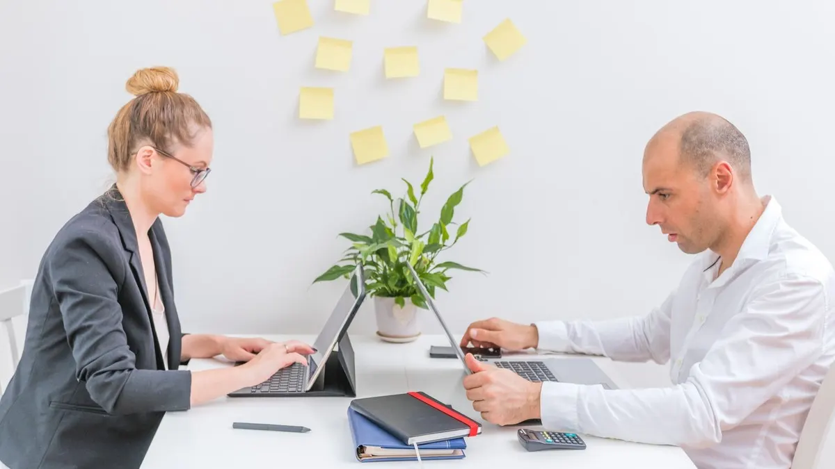 A woman and a man working on laptops across a white desk beneath a wall of yellow sticky notes in a bright office
