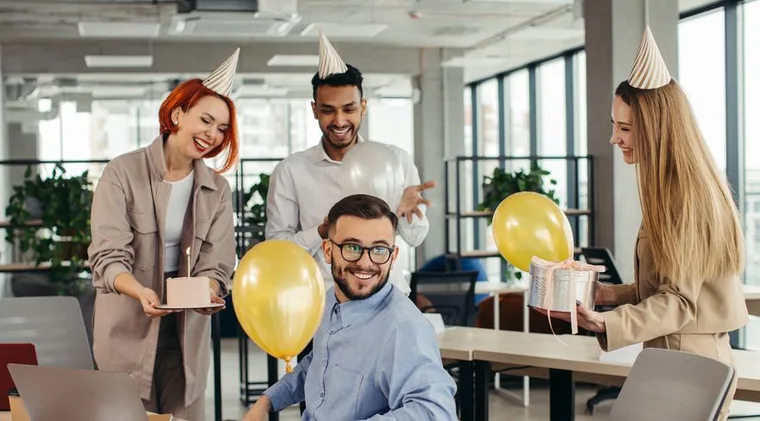 Colleagues in party hats surprising a teammate with cake and balloons at his desk