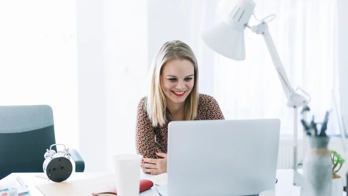 A smiling woman working on her laptop at a white desk in a bright, airy office