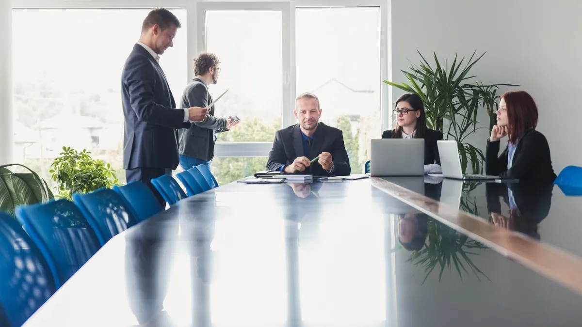 Five colleagues meet around a long glossy boardroom table beside bright windows