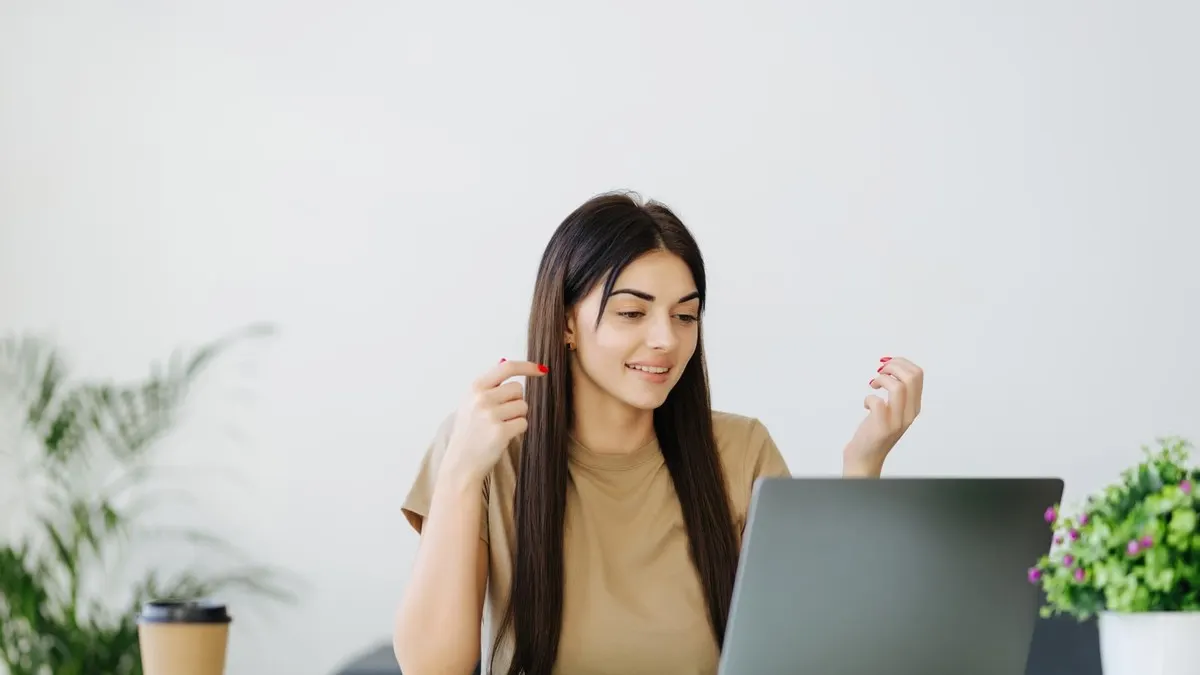 A smiling young woman raises her hands in excitement as she reads something on her laptop in a bright room.