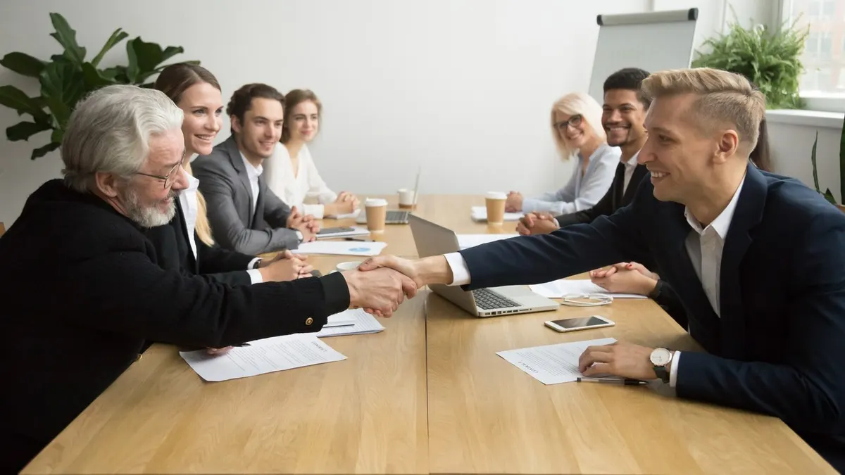An older and a younger businessman shaking hands across a meeting table as their colleagues look on in a bright white office
