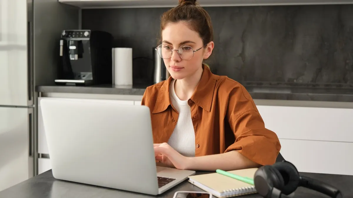 A young woman in round glasses studies on a laptop at a kitchen table with a notebook beside her