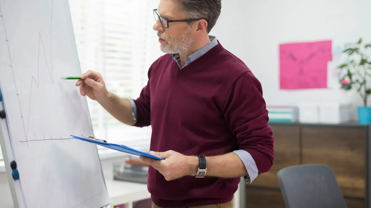 A grey-haired architect in glasses sketching a design on a flip chart with a clipboard in hand in a bright white office