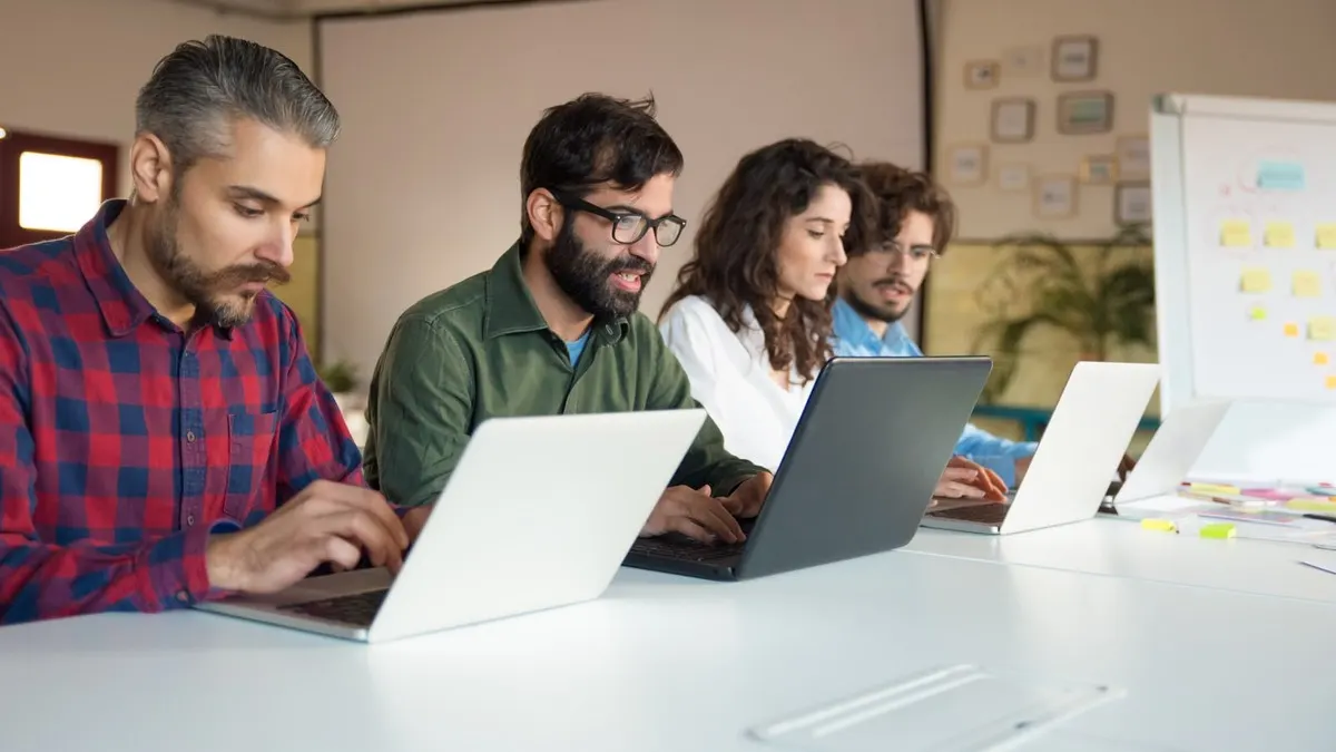 Four colleagues working side by side on their own laptops along a long white table in a bright office