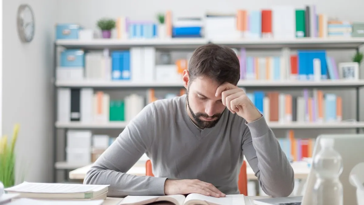 A man with his hand to his forehead concentrating on his study notes at a desk in a bright library