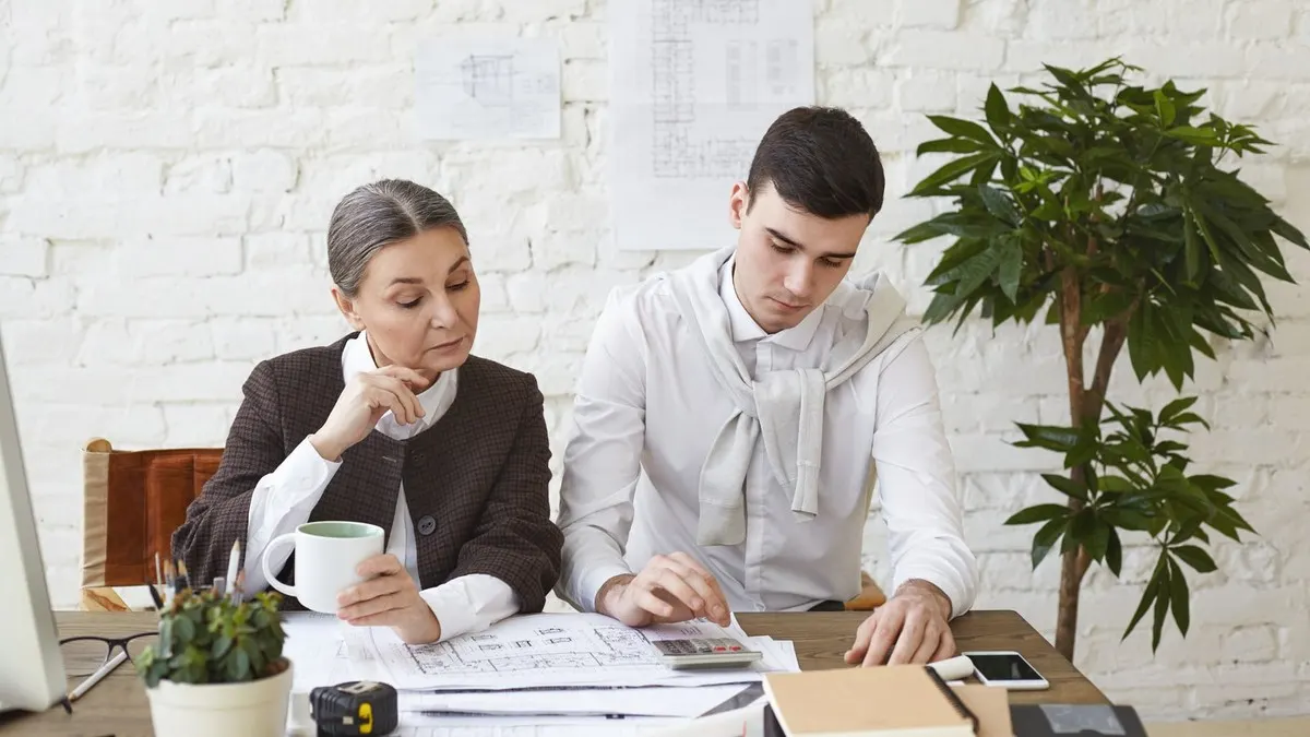 An architect works out figures on a calculator while a senior colleague studies the drawings beside him.