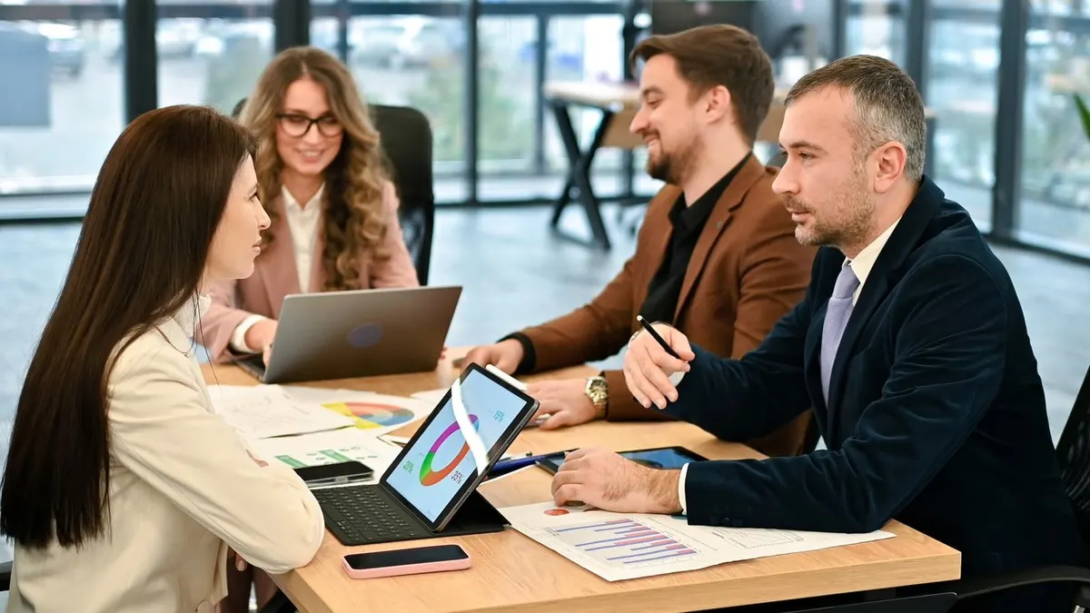 A marketing team reviewing charts on a tablet and laptops around a table in a bright office with tall windows