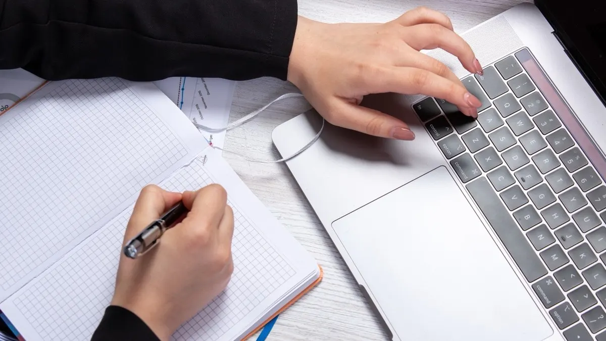 Hands writing revision notes in a grid notebook beside a laptop, with charts on the papers alongside