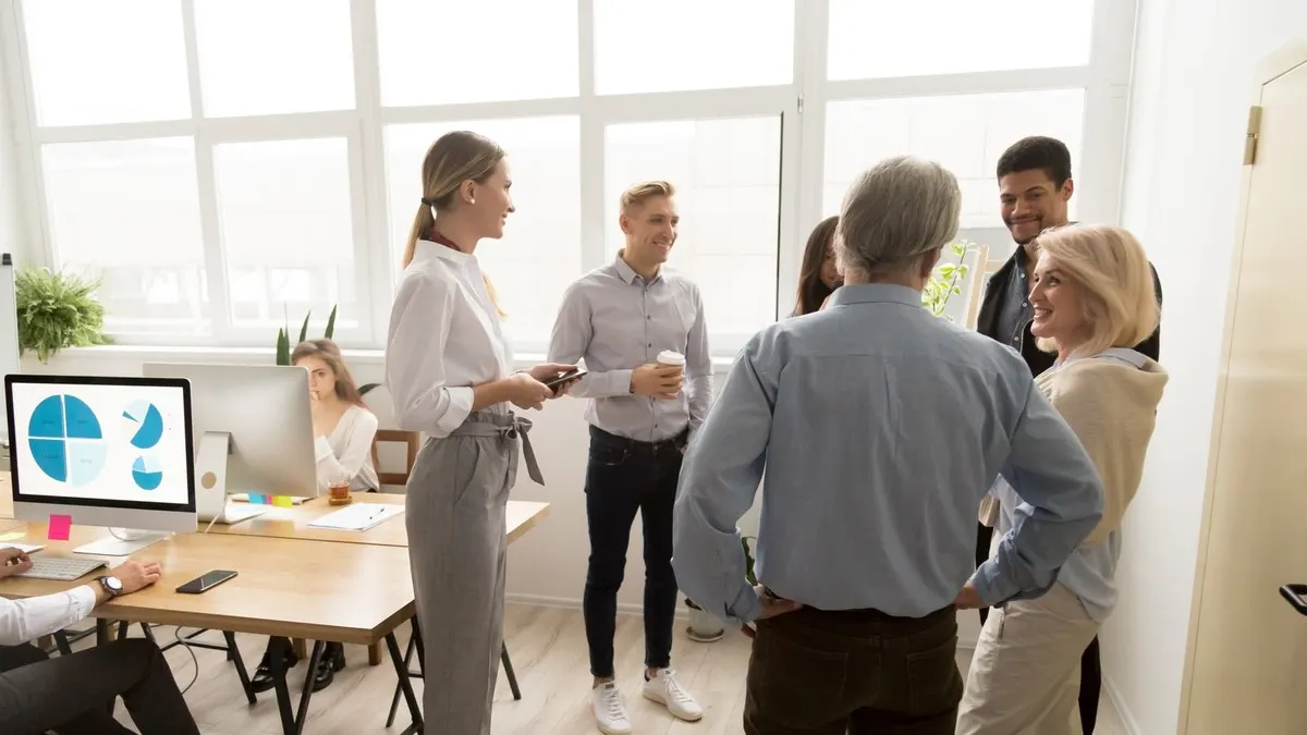 A team of young and senior colleagues standing together in a short huddle in a bright office with large windows