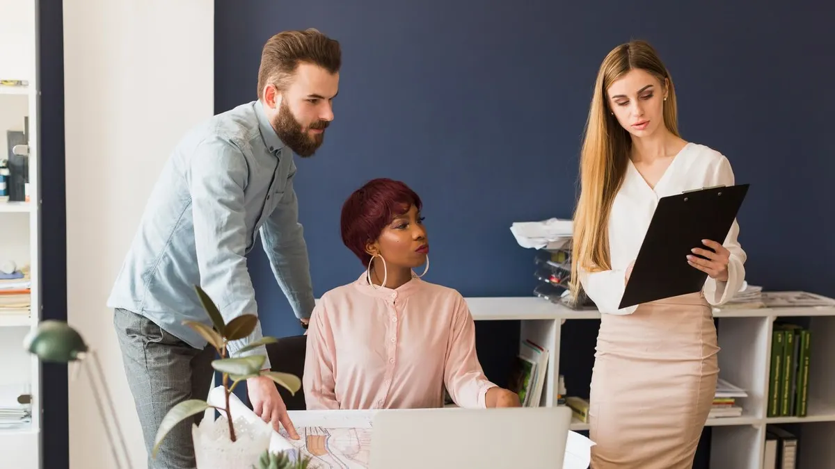 A woman reading from a clipboard while two colleagues look over plans at a desk in an office