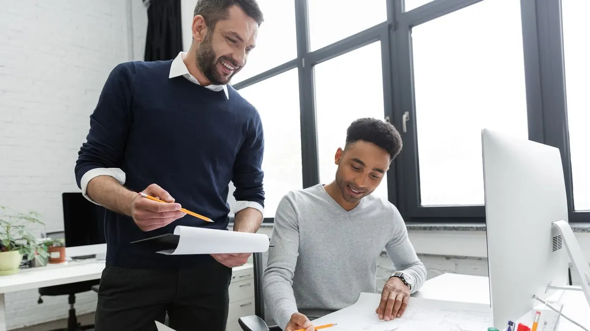 A smiling engineer holding design sketches talks through them with a colleague at his desk in a bright office with tall windows