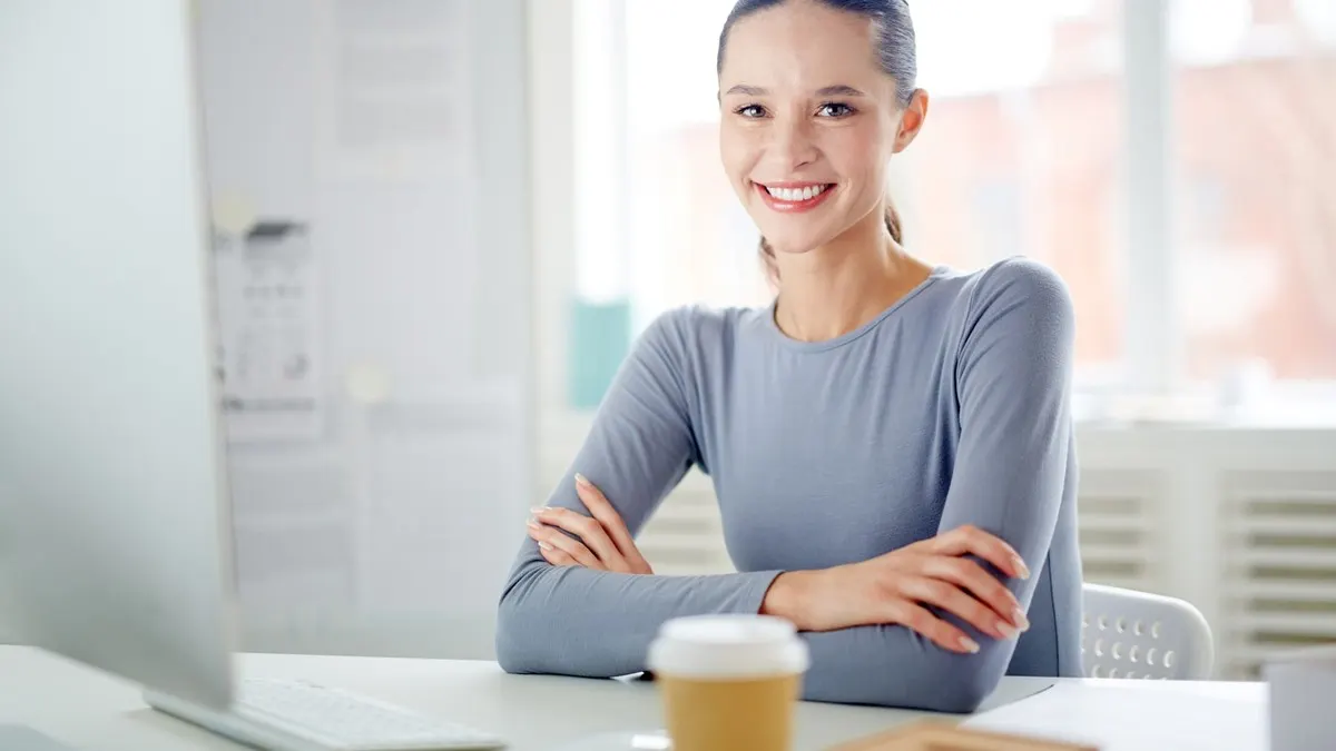 A smiling professional woman with folded arms sitting at her desk in a bright white office